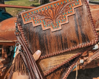 Könnte beinhalten: Eine braune und schwarze Kuhfell-Clutch mit einer geprägten Ledertasche mit einem türkisfarbenen Blumenmuster und türkisfarbenen Nieten. Die Clutch hat einen braunen Lederriemen mit einem Quasten.