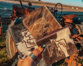 May include: Two brown and white cowhide zippered pouches with a brown leather zipper pull. The pouches have a brown embroidered design on the front. The pouches are held in a hand.