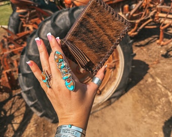 Op de afbeelding: Een bruine leren portemonnee met een kwastje. De portemonnee wordt vastgehouden in een hand die zilveren ringen en armbanden draagt met turquoise stenen. De armbanden hebben gegraveerde tekst die "Sage", "Prayers Up", "Down Deep" en "Work Baby" luidt.