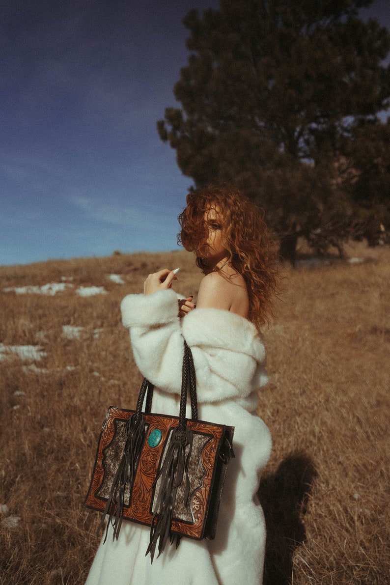 May include: A woman wearing a white fur coat holds a brown leather handbag with turquoise accents and fringe details. The bag has intricate tooling and a southwestern style.