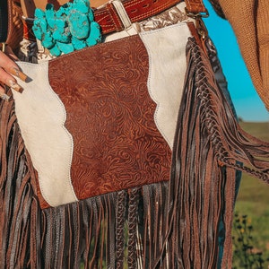 May include: A brown and white leather bag with fringe detailing. The bag has a southwestern style design and is decorated with turquoise stones.