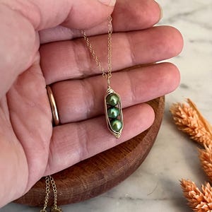 May include: A gold-colored necklace featuring a pendant shaped like a pea pod. The pea pod holds three green pearls. The necklace is held in a hand, with a wooden bowl and dried flowers in the background.