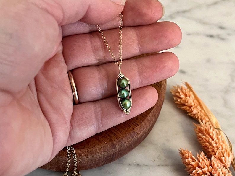 May include: A gold-colored necklace featuring a pendant shaped like a pea pod. The pea pod holds three green pearls. The necklace is held in a hand, with a wooden bowl and dried flowers in the background.
