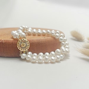 May include: A double strand pearl bracelet with a gold clasp featuring a floral design. The bracelet is displayed on a wooden surface.