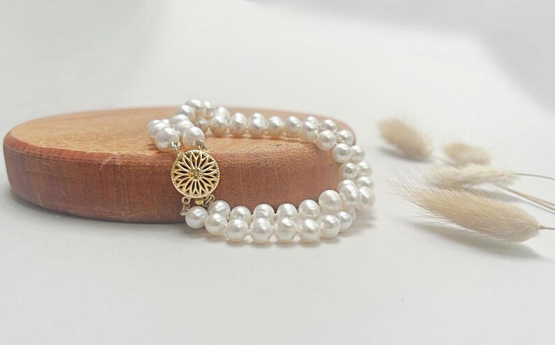 May include: A double strand pearl bracelet with a gold clasp featuring a floral design. The bracelet is displayed on a wooden surface.