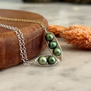 May include: Two pea pod necklaces, one gold-toned and one silver-toned, each holding three green pearls. The necklaces rest on a wooden surface with a marble background and a dried orange flower.