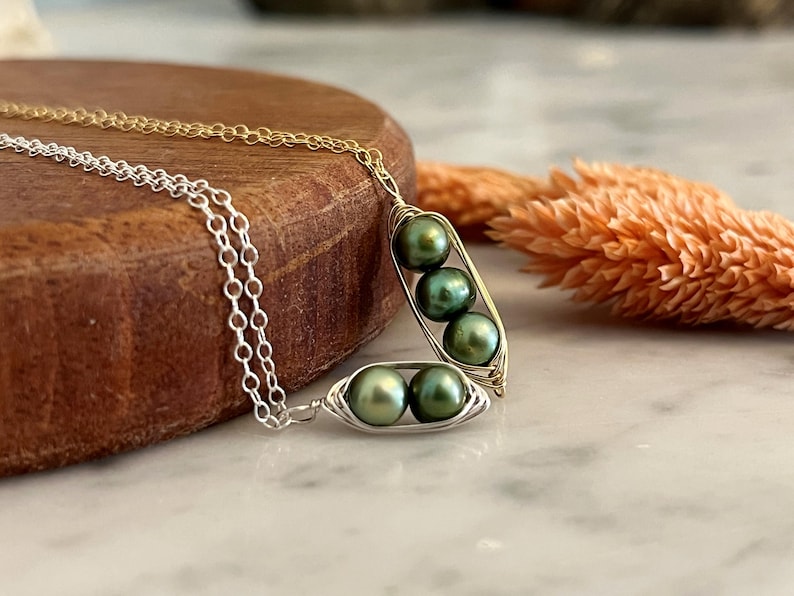 May include: Two pea pod necklaces, one gold-toned and one silver-toned, each holding three green pearls. The necklaces rest on a wooden surface with a marble background and a dried orange flower.