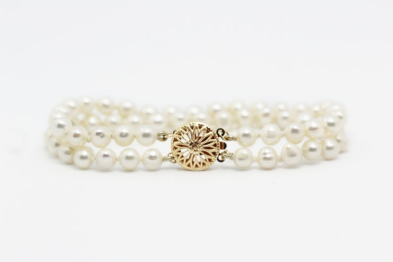 May include: A double strand pearl bracelet with a gold clasp. The clasp is a round, ornate design.