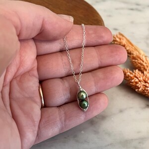 May include: A silver necklace featuring a pea pod pendant. The pendant holds two dark green pearls. The necklace is held in a hand, with a gold ring on the finger. Dried wheat stalks are in the background.