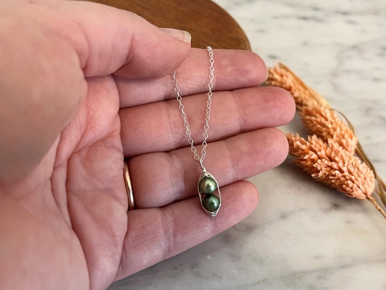 May include: A silver necklace featuring a pea pod pendant. The pendant holds two dark green pearls. The necklace is held in a hand, with a gold ring on the finger. Dried wheat stalks are in the background.
