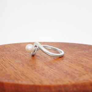 May include: A silver ring with a white pearl set in the band. The ring is curved and has a modern design.