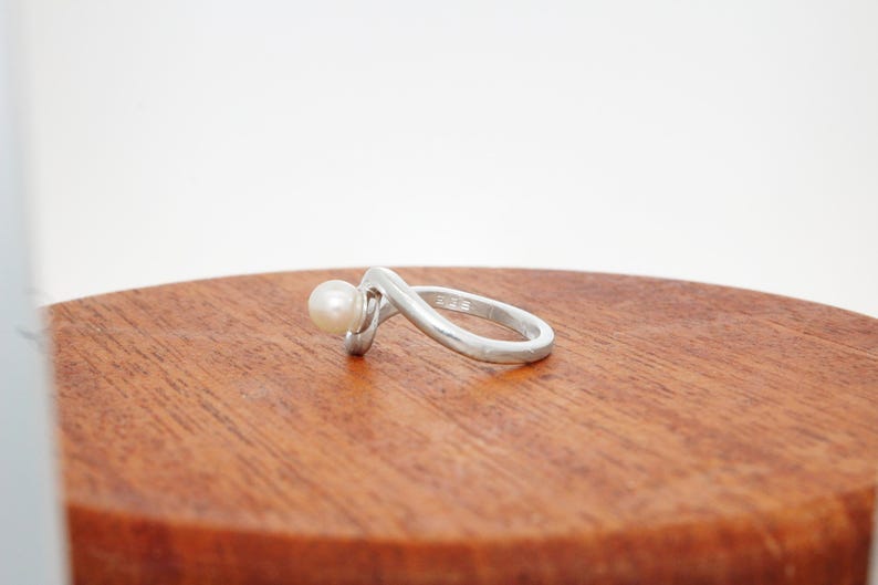 May include: A silver ring with a white pearl set in the band. The ring is curved and has a modern design.