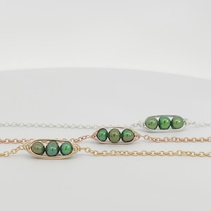 May include: Three bracelets featuring a pea-pod design, each containing three small, round, dark green beads. The bracelets are crafted from gold, rose gold, and silver-toned metal chains, with wire-wrapped frames holding the green beads.
