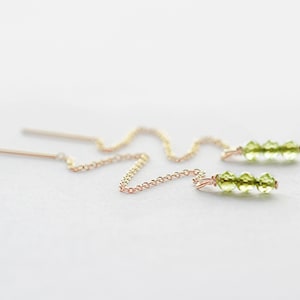 May include: A pair of gold chain earrings with green gemstone accents. The earrings are delicate and feature a small chain that hangs from the earlobe.