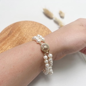 May include: A double strand pearl bracelet with a gold clasp. The bracelet is on a person's wrist.