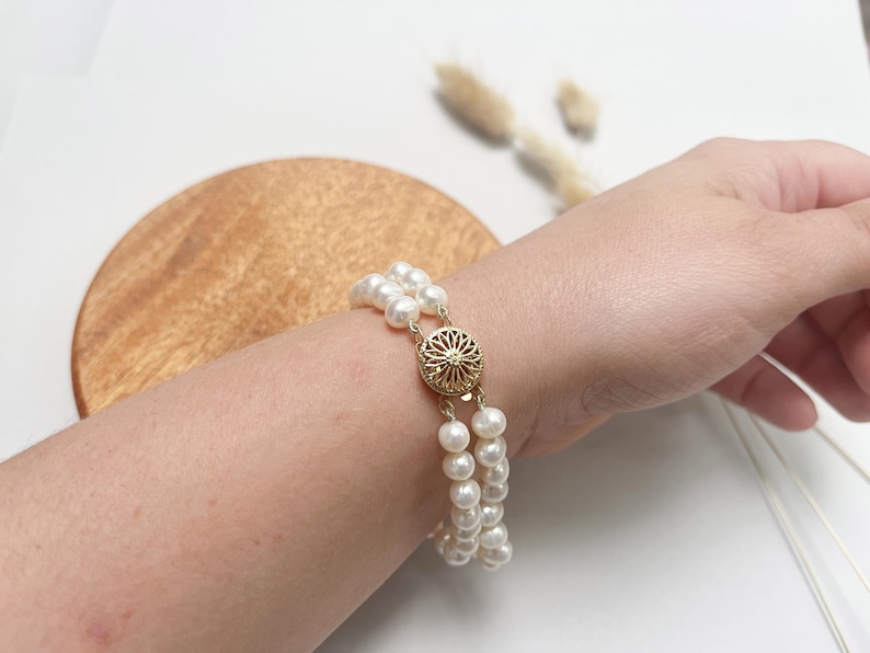 May include: A double strand pearl bracelet with a gold clasp. The bracelet is on a person's wrist.