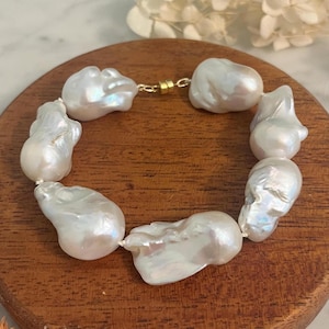 May include: A white pearl bracelet with a gold clasp. The bracelet features large, irregularly shaped pearls strung together. The pearls have a lustrous, iridescent sheen, reflecting light with a soft glow. The bracelet is displayed on a wooden surface.