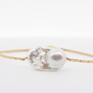 May include: A gold-toned bracelet featuring a large, irregularly shaped white pearl at its center. The bracelet's band has a textured finish. The pearl has iridescent hues, adding visual interest to this jewelry piece.