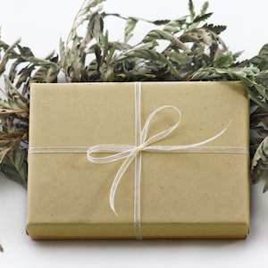 May include: A brown gift box with a white ribbon tied in a bow. The box is surrounded by dried foliage.