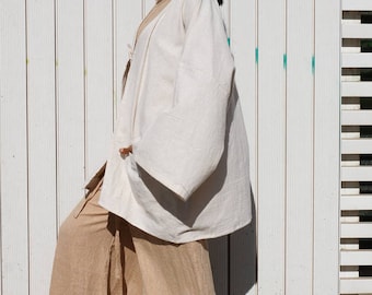 Puede incluir: Una mujer lleva un kimono de lino blanco sobre un pantalón de lino beige. El kimono tiene mangas anchas y un corte holgado. La mujer está de pie frente a una pared de madera blanca.