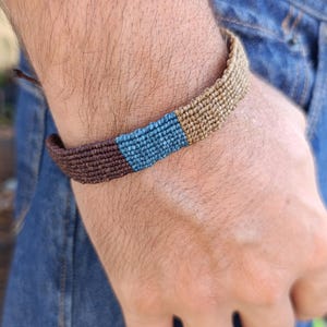 Könnte beinhalten: Ein gewebtes Armband mit einem Farbblock-Design. Das Armband hat ein braunes, blaues und beiges Farbschema. Das Armband wird am Handgelenk getragen, mit einer blauen Jeans im Hintergrund.