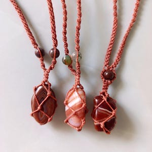 Puede incluir: Tres collares de piedras preciosas con cordones de macramé marrón rojizo. Cada collar presenta una piedra de diferente forma, con variaciones de color y patrón. Las piedras están encerradas en una red tejida, y los cordones tienen cuentas decorativas.