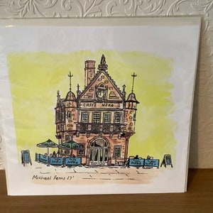 May include: A watercolor illustration of a building with a sign that reads "Caffe Nero". The building is a light brown color with a dark brown roof. There are people sitting outside the building at tables. The illustration is signed "Michael Ferns 17".