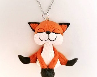 Op de afbeelding: Een handgemaakte ketting van polymer clay met een schattige oranje vos met gesloten ogen, zittend in een yogapose. De vos heeft zwarte ogen, een witte buik en zwarte voeten. De ketting is aan een zilveren ketting.