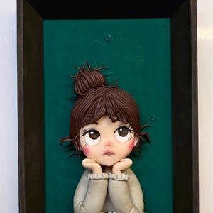 Puede incluir: Escultura enmarcada de una niña con cabello castaño recogido en un moño, apoyando la barbilla en sus manos. Lleva un suéter gris y está sentada frente a un fondo verde. Un libro abierto está frente a ella.