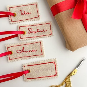 Four fabric gift tags with red stitching and gold eyelets. The tags have the names 'Isla', 'Sophie', and 'Anna' embroidered in red cursive. The tags are on a white surface with a red ribbon and a pair of gold scissors.