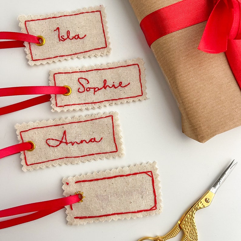 Four fabric gift tags with red stitching and gold eyelets. The tags have the names 'Isla', 'Sophie', and 'Anna' embroidered in red cursive. The tags are on a white surface with a red ribbon and a pair of gold scissors.