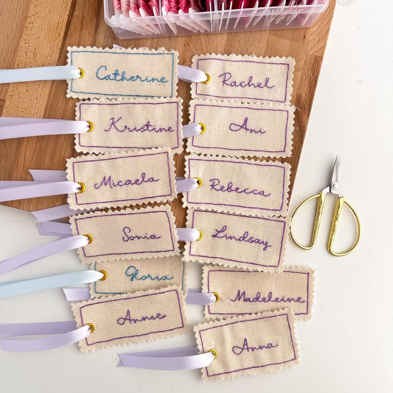 A collection of 12 beige fabric tags with purple, blue, and gold embroidery. The tags are decorated with a ribbon and have names embroidered on them: Catherine, Rachel, Kristine, Ann, Micaela, Rebecca, Sonia, Lindsay, Gloria, Madeleine, Annie, and Anna.