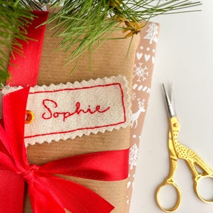 A brown wrapped paper gift  with a red ribbon tied around it. A white fabric tag with red stitching is attached to the bag with the name 'Sophie' embroidered in red thread. A pair of gold scissors are on the right side of the image.