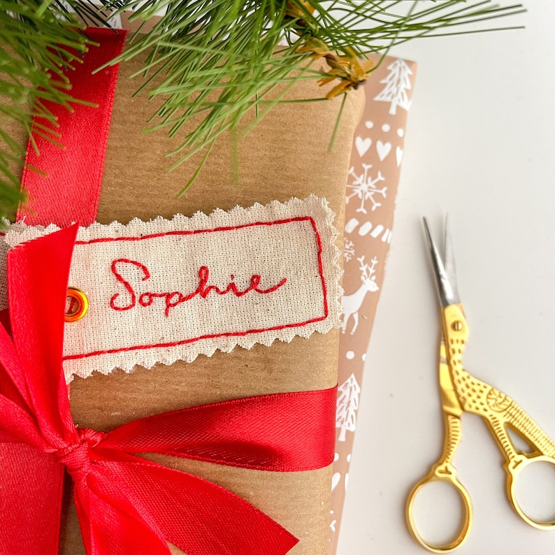 A brown wrapped paper gift  with a red ribbon tied around it. A white fabric tag with red stitching is attached to the bag with the name 'Sophie' embroidered in red thread. A pair of gold scissors are on the right side of the image.