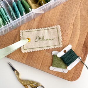 A beige fabric tag with a scalloped edge and a gold eyelet. The tag is embroidered with the name 'Ethan' in green thread. The tag is tied to a light green ribbon. Two spools of green embroidery floss are visible in the background.