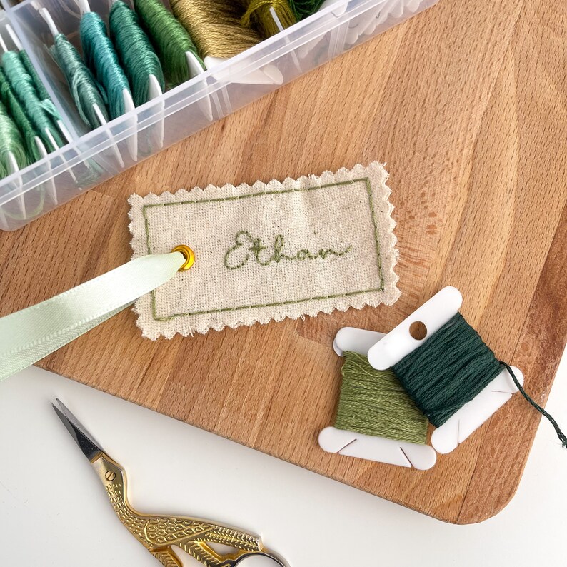 A beige fabric tag with a scalloped edge and a gold eyelet. The tag is embroidered with the name 'Ethan' in green thread. The tag is tied to a light green ribbon. Two spools of green embroidery floss are visible in the background.