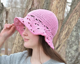 May include: A light pink crochet bucket hat featuring a textured, dense stitch pattern on the crown and a wide, scalloped, open-work brim with a delicate floral-inspired design that flares outward from the base of the hat.