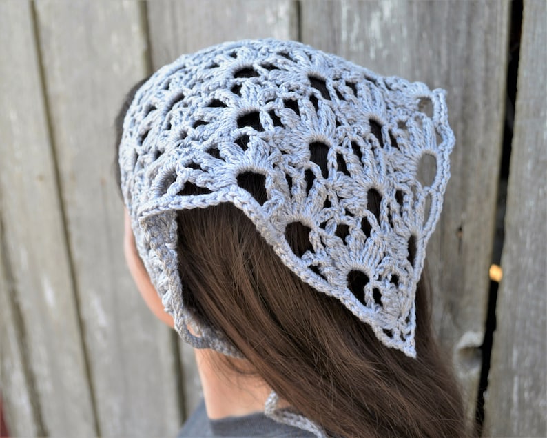 May include: A light gray crocheted bandana with a delicate floral pattern. The bandana is tied around the head and hangs down the back.