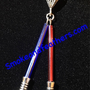 May include: A silver necklace with a triangular pendant and two dangling light blue and red lightsaber-shaped charms. The charms have silver caps and black tips. The necklace is on a black background.