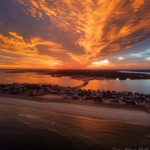 May include: Aerial view of a coastal town at sunset with a bridge over a body of water. The sky is a vibrant orange and pink with clouds. The water reflects the colors of the sky.