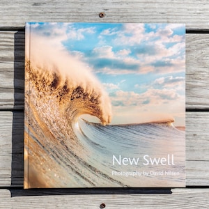 May include: A square book titled "New Swell" featuring a photograph of a large ocean wave curling over. The wave is a mix of gold, white, and blue, with a cloudy sky in the background. The book's cover also includes the photographer's name.