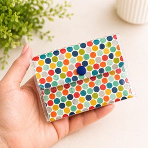 May include: A small, clear plastic wallet with a colorful polka dot pattern. The pattern features various colors, including red, orange, yellow, green, and blue. The wallet is secured with a blue button closure.