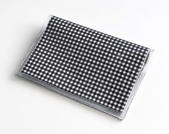 Puede incluir: Un tarjetero o billetera rectangular con un patrón clásico de cuadros vichy en blanco y negro. El artículo está recubierto por una funda de plástico transparente que proporciona una capa protectora sobre el material.