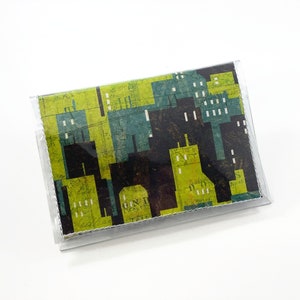 May include: A rectangular card holder with a cityscape design. The design features black, teal, and lime green buildings. The card holder is encased in a clear plastic sleeve.