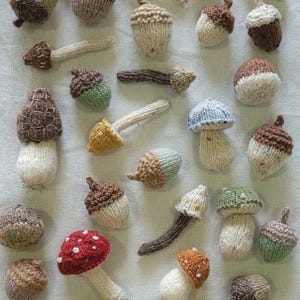 Puede incluir: Una colección de setas y bellotas tejidas a mano en varios colores. Las setas tienen sombreros rojos, naranjas, amarillos, azules y verdes con tallos blancos. Las bellotas son marrones y verdes. Los artículos están dispuestos sobre una superficie blanca.