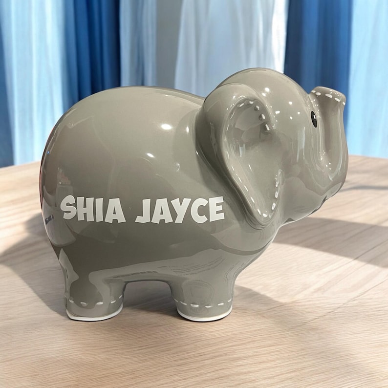 May include: A gray ceramic elephant piggy bank with the name "SHIA JAYCE" written in white on the side.