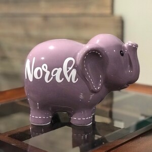 May include: Purple ceramic elephant piggy bank with the name "Norah" written in white on the side.