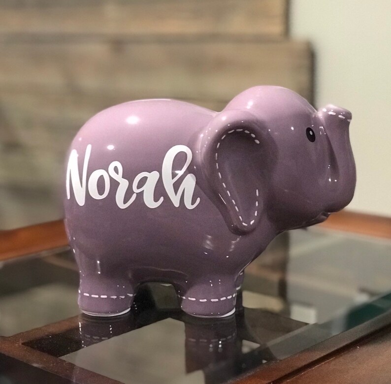 May include: Purple ceramic elephant piggy bank with the name "Norah" written in white on the side.