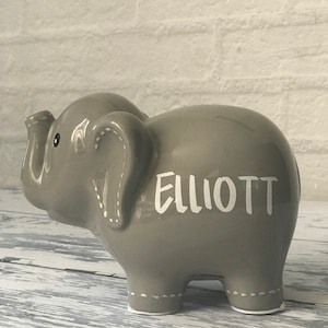 May include: Gray ceramic elephant piggy bank with white stitching detail and the name "ELLIOTT" written in white on the side.