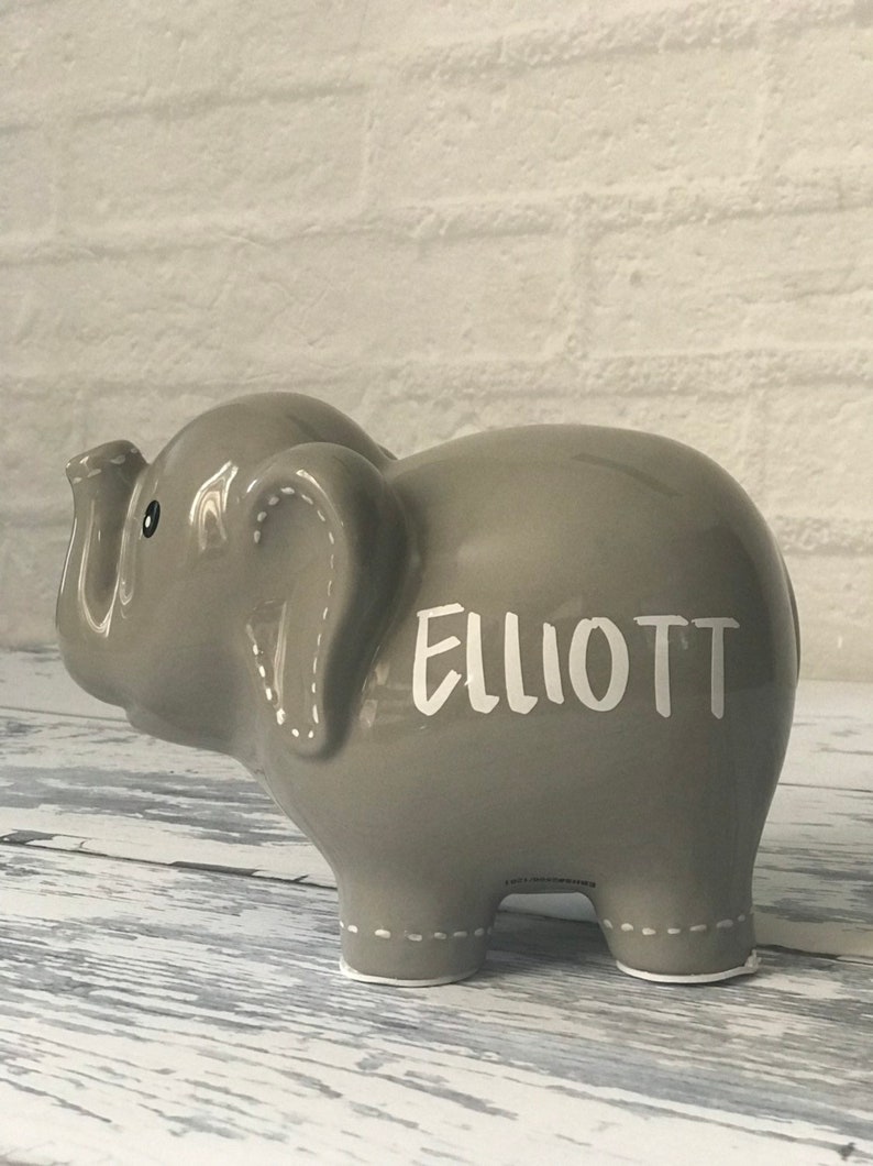 May include: Gray ceramic elephant piggy bank with white stitching detail and the name "ELLIOTT" written in white on the side.